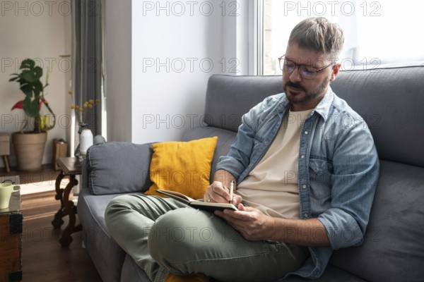 A man with glasses sits on a comfortable sofa, writing in a notebook with a pen. Sunlight streams through the window, casting a warm glow in the cozy living room