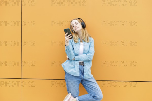 A stylish woman leans against an orange wall in Warsaw, wearing headphones and checking her phone, enjoying the vibe of urban technology and modern style