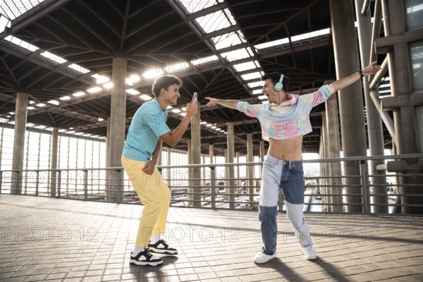 A lively gay couple enjoys a playful moment in an urban setting. One person, wearing colorful attire, dances energetically while the other captures the moment with a smartphone