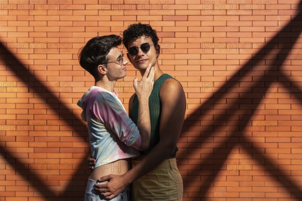 A gay couple shares an affectionate embrace against a sunlit brick wall, casting interesting shadows. Both wear casual, colorful outfits and sunglasses, exuding warmth and love