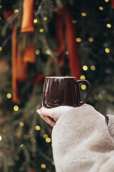 Cropped unrecognizable young woman holding a dark brown mug in front of a decorated Christmas tree with shimmering lights. The winter atmosphere is evoked by her warm clothing and the festive background