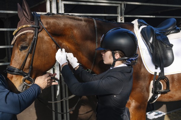 A teenager in classical dressage attire adjusts a horse's bridle with assistance, focusing intently on the task. Both the horse and the rider are well-prepared for training
