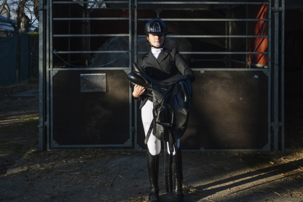 A teenage girl in classical dressage attire confidently holds a saddle. She stands by a stable, looking directly at the camera