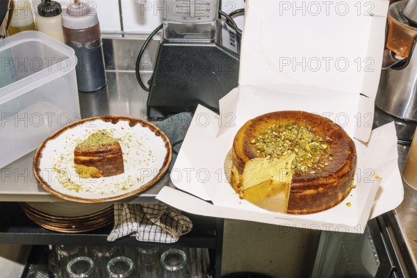 A rich cheesecake topped with crushed pistachios sits in an open box, with a slice served on a plate nearby. The scene is set in a coffee shop kitchen with baking tools and ingredients