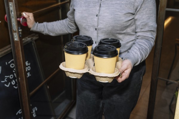 A person opens a cafe door while holding a tray of four takeaway coffee cups. The setting suggests a warm, inviting atmosphere, emphasizing the specialty coffee theme
