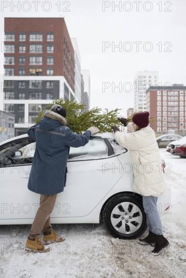 A couple wearing winter clothing secures a Christmas fir onto their car's roof in a snow-covered city setting Buildings in the background provide an urban holiday atmosphere