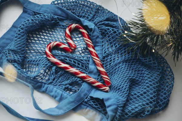 A vibrant blue string bag lays on a white surface, gently cradling two candy canes formed into a heart shape. Surrounded by soft holiday lights and pine branches, this scene captures a unique holiday spirit with a touch of sweetness
