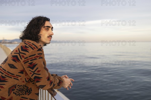 A man with a thoughtful expression leans against a railing, gazing at the tranquil sea The natural tones of his sweater complement the serene ocean horizon
