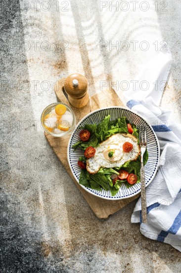 Top view of a nutritious and vibrant meal featuring fresh spinach leaves, cherry tomatoes, and a perfectly fried egg, paired with a refreshing mineral water with ice and kumquat slices, neatly presented on a rustic kitchen surface