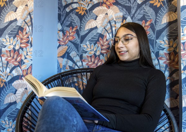 A young woman enjoys reading a book, seated in a modern chair against a colorful floral-patterned background