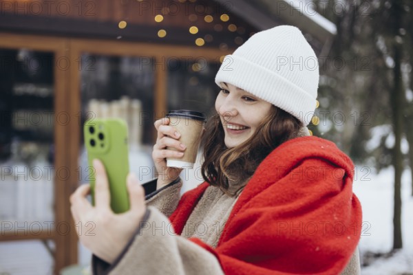 A cheerful young woman in a white beanie and red scarf smiles while holding a coffee cup and taking a selfie with a smartphone on a snowy winter day
