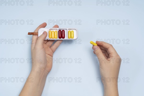 Top view of a cropped unrecognizable person's hand holding a white pill box filled with colorful capsules on a light blue background. A yellow pill is being held between fingertips, showcasing the concept of medication management