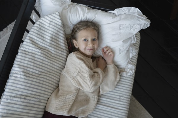 A child with light hair and blue eyes lies comfortably in bed, wearing a cozy sweater The bed is adorned with a striped pillow, creating a peaceful and relaxed atmosphere