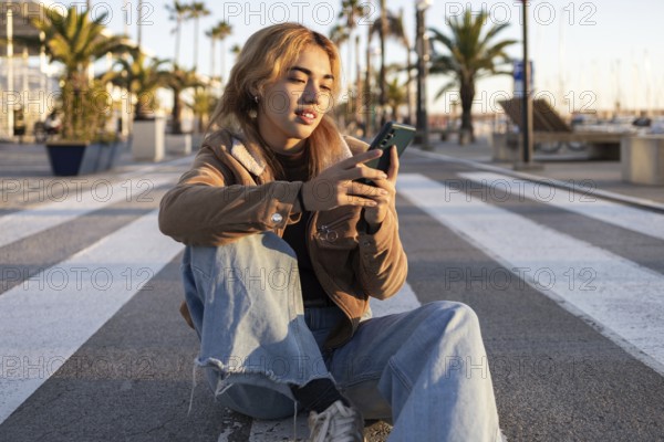 Mixed-race woman sits cross-legged on an outdoor boardwalk, absorbed in her smartphone Palm trees and a serene sunset create a relaxed, modern atmosphere