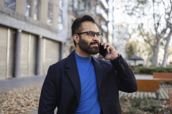 Indian businessman in winter clothing using a mobile phone in an urban park setting, looking away from the camera