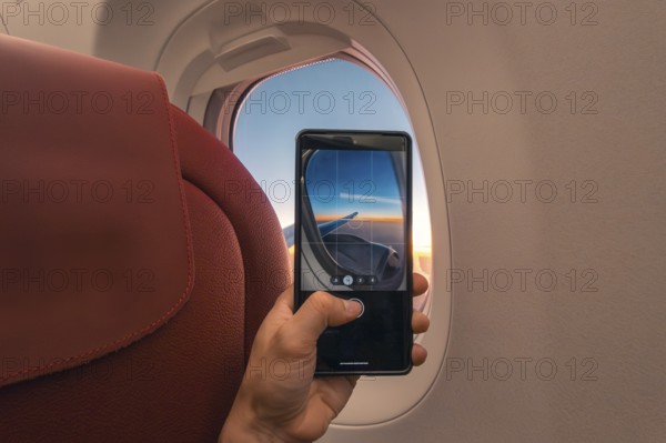 Cropped unrecognizable person captures a sunset over the ocean using a smartphone, viewed through an airplane window