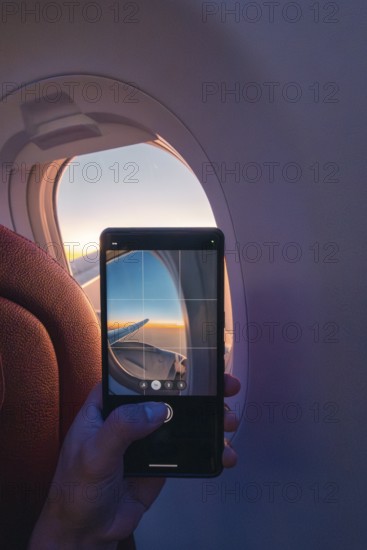 Cropped unrecognizable person capturing a serene sunset through an airplane window using a smartphone, highlighting the use of technology to record beautiful moments mid-flight