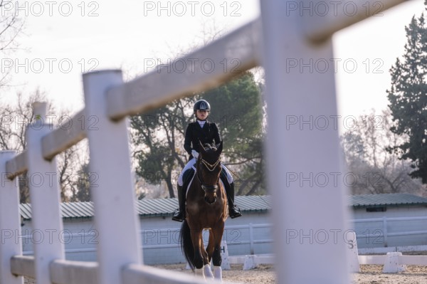 A teenage girl competently engages in classical dressage, riding a horse in an outdoor arena. Dressed traditionally in a black jacket and helmet, she directs her gaze forward, focused on her path