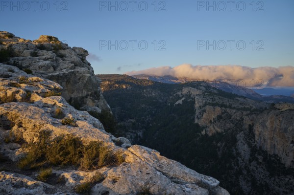 A stunning landscape of rocky cliffs and forested valleys illuminated by the warm light of sunset, with a cloud-covered horizon in the serene mountains of Alicante, Spain