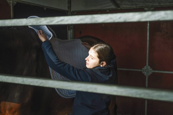 A teenage girl carefully places a saddle blanket on a horse in preparation for a classical dressage session, demonstrating focus and care inside a stable