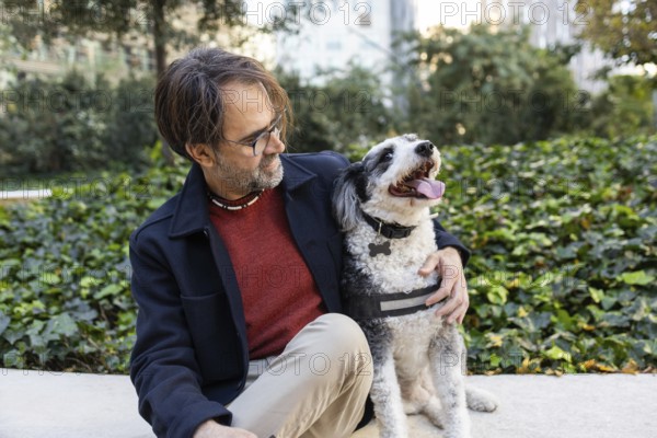 A middle-aged man interacts with his black and white dog in a city park, surrounded by lush greenery and modern buildings in the background. Both appear joyful and at ease in this urban setting