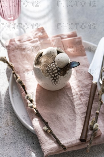 From above, an artistic Easter table arrangement features a porcelain bowl with speckled eggs, adorned with delicate feathers, beside a pastel pink napkin and elegant silverware under soft daylight