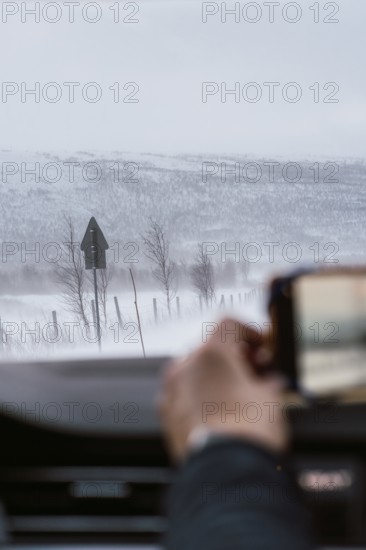 A hand captures a snowy landscape from inside a vehicle, showcasing the tranquil and harsh beauty of Swedish Lapland's winter roads, with snow-covered fields and distant mountains