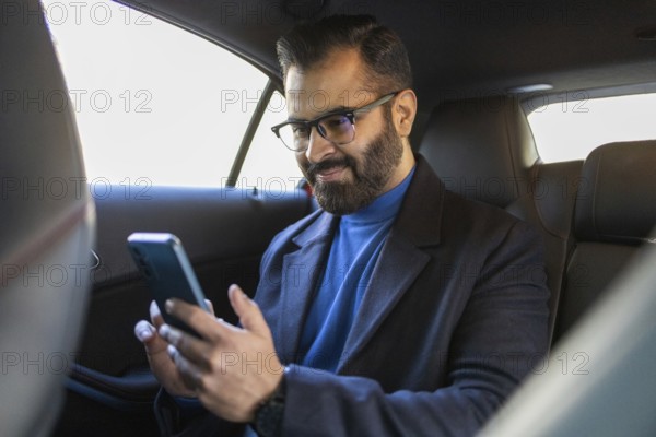 Indian man in blue turtleneck and black coat, using smartphone in car, looking at mobile screen