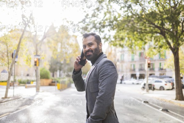 Cheerful Indian man in winter business attire, on a call using a mobile phone on a sunny urban street, embodying a modern, active lifestyle