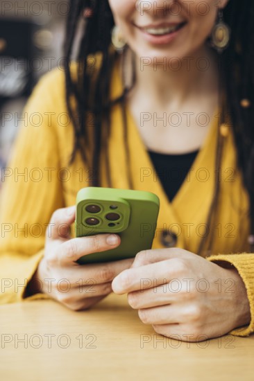 A woman in a mustard yellow sweater holds a smartphone with a green case, smiling The cozy setting and colors create a warm and inviting atmosphere