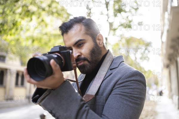An Indian man, dressed professionally in a grey coat, focuses intently on capturing a photo with his advanced camera on a sunny city street