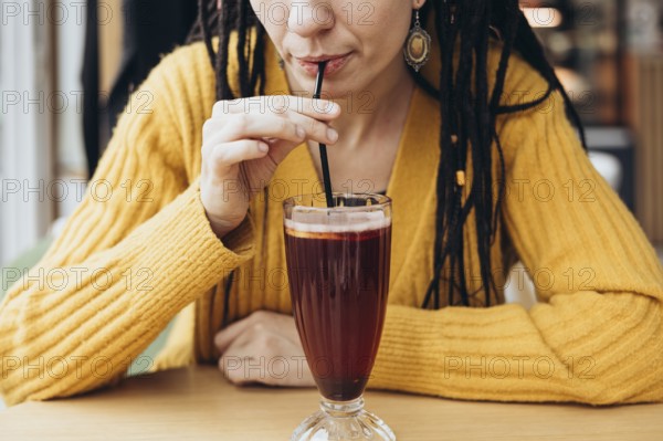 A woman in a yellow sweater sips a refreshing coffee drink through a straw in a cozy cafe setting The focus is on her hands and the drink, creating a warm ambiance