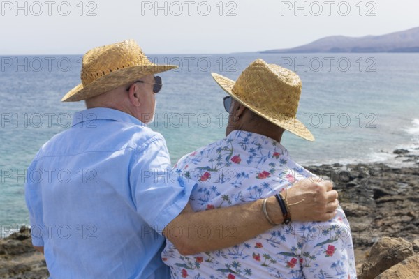 Back view of an unrecognizable senior gay couple standing close together, looking out towards the sea. Both men wear straw hats and casual shirts, embodying a moment of peaceful retirement by the ocean