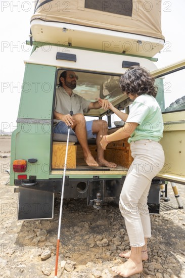 A blind man sits at the back of a motorhome, holding a cane, while a supportive woman assists him. They embrace the joy of adventure and inclusivity in outdoor exploration