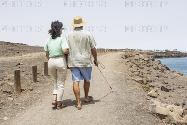 A blind man walking with a female guide along a coastal path. He uses a cane to navigate the rocky terrain, embracing the motorhome lifestyle in a natural environment