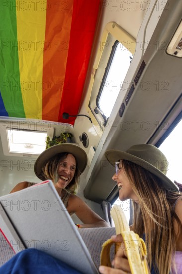 Lesbian couple laughing inside a van decorated with a rainbow flag. One holds a book while the other eats a banana, capturing a joyful, spontaneous moment on the road