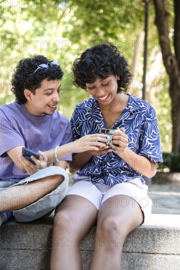 A cheerful multiethnic LGBTQ+ couple sitting outdoors, sharing and looking at a phone together, portraying happiness, diversity, and love in a sunny park setting