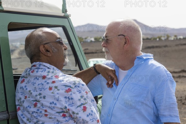 A senior gay couple shares a tender moment beside their off-road vehicle during a camping trip. Both men gaze at each other lovingly, emphasizing a warm, affectionate connection in a serene natural setting