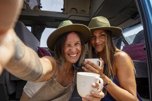 Lesbian couple wearing hats enjoy coffee inside a camper van, capturing a joyful moment of adventure and travel. The scene conveys freedom on the road