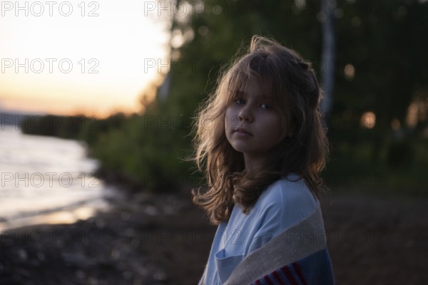 A young girl with wavy hair gazes thoughtfully at the camera while standing by a lakeside at sunset She is wrapped in a cozy blanket, evoking a calm, serene mood