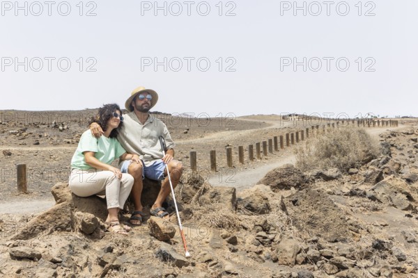 A blind man with a cane and a woman sit together on rocks along a quiet desert path. The scene captures a moment of rest and companionship during their outdoor exploration