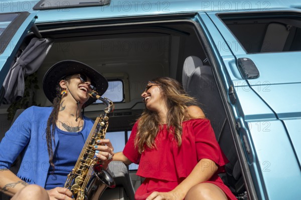 A joyful lesbian couple smiles while relaxing by their van. One plays a saxophone, capturing the essence of freedom and love during a sunny summer adventure