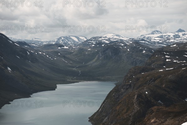 A sweeping view of a tranquil lake nestled in a dramatic mountain valley in Norway Snow-dusted peaks and cloudy skies frame this breathtaking Nordic wilderness scene
