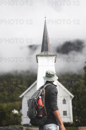 A traveler with a backpack gazes at a quaint white church amid Norway's majestic landscape Misty mountains envelop the scene, evoking adventure, exploration, and tranquility