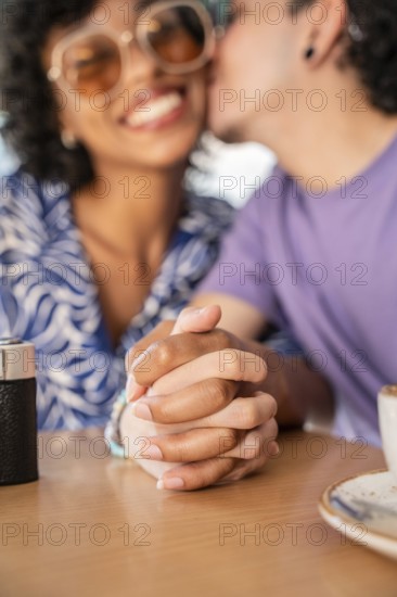 A cheerful multiethnic LGBTQ+ couple seated at a table, holding hands and sharing a tender moment, symbolizing love and diversity