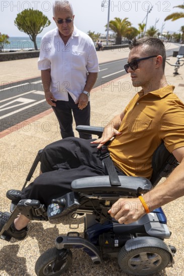 An older man and a younger man in a wheelchair share a moment outdoors on a sunlit pathway by the sea. The older man, looks attentively at the younger man, who is dressed in a mustard polo shirt and navigates an electric wheelchair