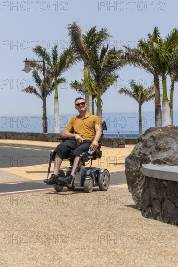 A man in a wheelchair is captured enjoying the sunny weather on a paved path. He is dressed in a casual yellow shirt and black shorts, with sunglasses on, exuding a relaxed vibe amidst tropical palm trees and clear skies