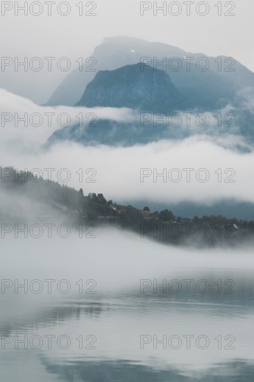 A peaceful lake reflects layers of mist and towering mountains in Norway The hazy atmosphere and soft light create a serene and dreamy Nordic landscape