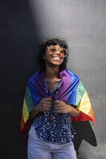 A smiling LGTBQ+ person wearing sunglasses and a patterned shirt, joyfully wrapped in a rainbow flag, embodies pride and happiness against a neutral background with sunlight
