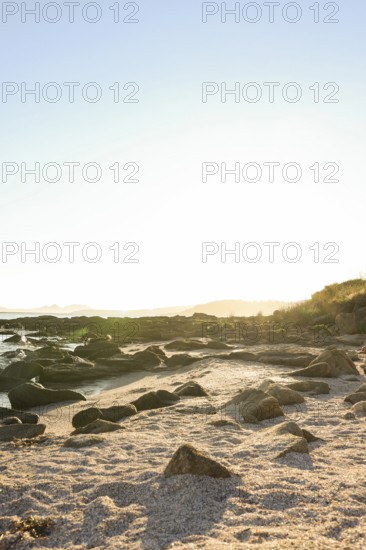 A tranquil coastal scene at sunrise, displaying golden sunlight illuminating a rocky beach The calm ocean and sandy foreground create a peaceful atmosphere, ideal for nature and relaxation themes
